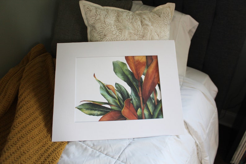 May include: A framed watercolor painting of a leafy plant with green and orange leaves. The painting is on a white background and is framed in a white frame.