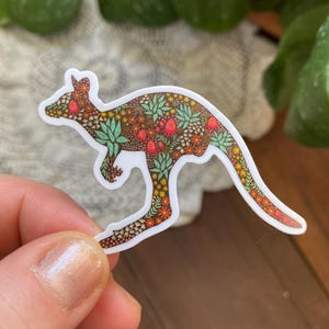 Peut inclure: Un autocollant représentant un kangourou avec un motif floral. Le kangourou est brun avec des fleurs rouges, orange, jaunes et vertes. L'autocollant est sur un fond blanc.