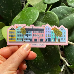 Op de afbeelding: Een sticker met een rij kleurrijke huizen met palmbomen ervoor. De sticker is roze en heeft onderaan de tekst "Charleston, SC".