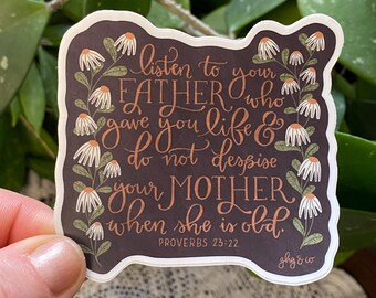 May include: A dark gray sticker with white daisy flowers around the edges. The sticker features a quote in brown cursive lettering: "Listen to your father who gave you life & do not despise your mother when she is old." The quote is attributed to Proverbs 23:22. The sticker also includes the text "ghg & co" in brown cursive lettering.