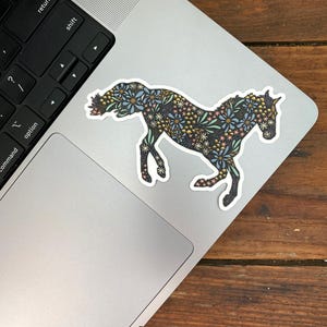May include: A medium-sized sticker of a horse in a running pose, decorated with a floral pattern in shades of blue, yellow, and pink. The sticker is placed on a laptop. The size is 4" x 2.92".