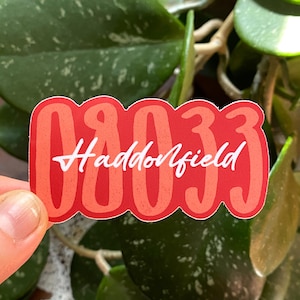 May include: A red and coral sticker with the text "08033 Haddonfield" in a bold, rounded font. The sticker is held in front of a background of green leaves. The bottom of the sticker reads "RED/CORAL".