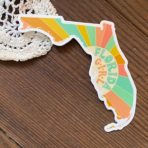 May include: A sticker of the state of Florida with a retro sunburst design and the text "Florida Girl" in a circle.