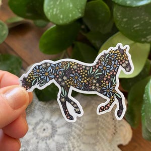 May include: A sticker of a horse in a running pose, decorated with a floral pattern in shades of blue, yellow, and pink on a black background. The sticker is approximately 3 inches by 2.19 inches.