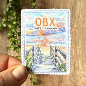 May include: A rectangular sticker featuring a watercolor painting of a beach boardwalk leading to the ocean under a colorful sunset. The sticker has the text "OBX NORTH CAROLINA" in orange and blue.
