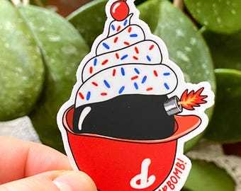 May include: A sticker featuring a cupcake design with a bomb as the base, topped with white frosting and red, white, and blue sprinkles. A red cherry tops the frosting. The cupcake sits in a red cup, with the text "SCHWARBOMB!" in white.