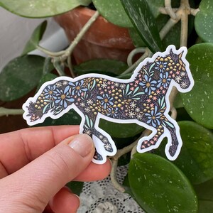 May include: A sticker in the shape of a horse, decorated with a floral pattern in shades of blue, pink, and gold on a black background. The sticker is held by a hand. The text at the bottom reads: SIZE PICTURED: MEDIUM: 4" x 2.92".