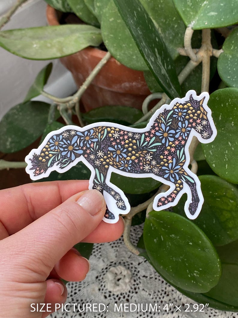 May include: A sticker in the shape of a horse, decorated with a floral pattern in shades of blue, pink, and gold on a black background. The sticker is held by a hand. The text at the bottom reads: SIZE PICTURED: MEDIUM: 4" x 2.92".