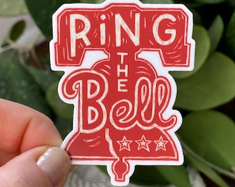 May include: A red and white sticker with the words "RING THE BELL" in a vintage style. The design includes a bell shape with three stars at the bottom. The sticker is held by a person's hand against a background of green leaves.