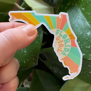 May include: A sticker shaped like the state of Florida with a retro sunburst design in shades of orange, yellow, green, and blue. The text "FLORIDA GIRL" is written vertically in a white outline.