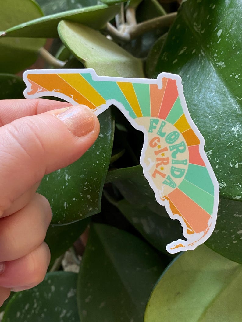 May include: A sticker shaped like the state of Florida with a retro sunburst design in shades of orange, yellow, green, and blue. The text "FLORIDA GIRL" is written vertically in a white outline.