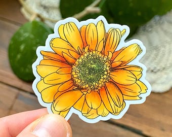 May include: A vibrant sticker featuring a watercolor illustration of a gerbera daisy. The flower has bright yellow and orange petals with a green center. The sticker is held by a hand, with a wooden surface and green foliage in the background.