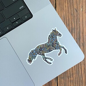 May include: A sticker of a horse in a running pose, decorated with a floral pattern in blue, white, and gold. The sticker is on a laptop. The text at the bottom reads: SIZE PICTURED: SMALL 3" X 2.19".