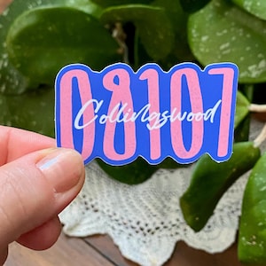 May include: A blue sticker with pink text that reads "08107" above the word "Collingswood" in white script. The sticker is held in front of a background of green leaves.
