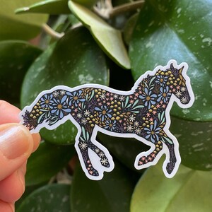 May include: A sticker featuring a horse silhouette filled with a floral pattern in blue, yellow, and white. The sticker is approximately 3 inches by 2.19 inches. The horse is depicted in a running pose, with a white border.