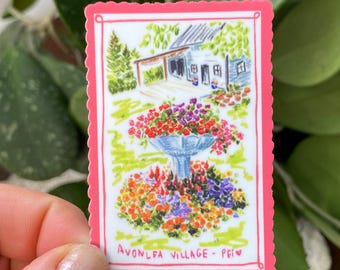 May include: A rectangular sticker with a pink border featuring a watercolor illustration of a blue fountain surrounded by colorful flowers. A house with a porch and trees is in the background. The text "AVONLEA VILLAGE - PEI" is at the bottom.