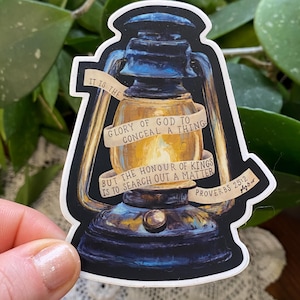 May include: A sticker featuring a detailed illustration of an antique lantern. The lantern is primarily black and gold, with a glowing interior. Text on the sticker reads, "IT IS THE GLORY OF GOD TO CONCEAL A THING" and "BUT THE HONOUR OF KINGS IS TO SEARCH OUT A MATTER PROVERBS 25:2".