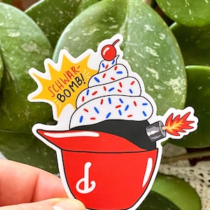 May include: A medium-sized sticker featuring a cupcake bomb design. The cupcake has white frosting with red, white, and blue sprinkles, topped with a cherry. The bomb base is black, with a red fuse and flames. The text "SCHWAR-BOMB!" is in a yellow burst. The sticker measures 3" x 3.14".