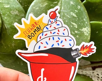 May include: A medium-sized sticker featuring a cupcake bomb design. The cupcake has white frosting with red, white, and blue sprinkles, topped with a cherry. The bomb base is black, with a red fuse and flames. The text "SCHWAR-BOMB!" is in a yellow burst. The sticker measures 3" x 3.14".