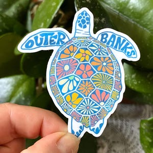 May include: A colourful sticker in the shape of a turtle, featuring the text "Outer Banks". The turtle's shell is decorated with a floral pattern in shades of blue, orange, yellow, and pink. The sticker measures 6.76 cm x 7.57 cm.
