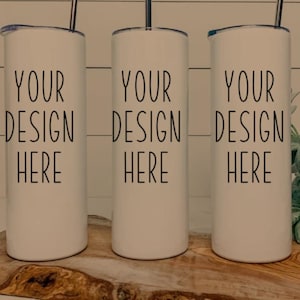 May include: Three white insulated tumblers with black text that reads "YOUR DESIGN HERE". Each tumbler has a silver straw and a lid. The tumblers are cylindrical and sit on a wooden surface.