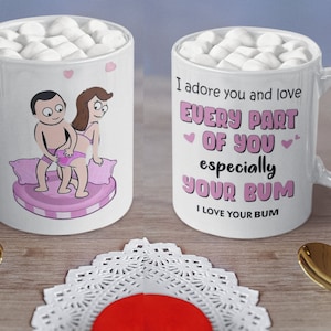 May include: Two white ceramic mugs filled with marshmallows. One mug features a cartoon couple in a romantic pose. The other mug has the text "I adore you and love EVERY PART OF YOU especially YOUR BUM".