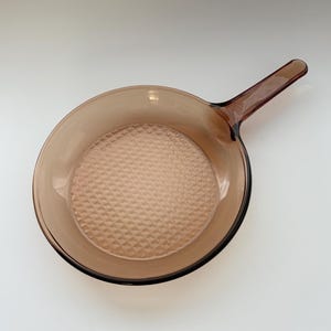 May include: A vintage brown glass skillet with a long handle. The interior of the pan has a textured, diamond-patterned surface. The skillet is made of transparent glass and is designed for cooking.
