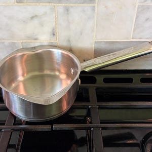 May include: A small, stainless steel saucepan with a long handle, resting on a black stovetop. The pan has a curved shape and a shiny surface. The background features a marble tile backsplash.