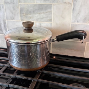 May include: A stainless steel saucepan with a lid and a black handle. The pot has a wooden-colored base and a wooden knob on the lid. It sits on a black stovetop with a marble tile backsplash.