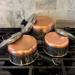 May include: A set of three stainless steel pots with copper-colored bases and black handles. Two pots have matching stainless steel lids with black knobs. The cookware is arranged on a black stovetop, showing metallic surfaces and circular designs.