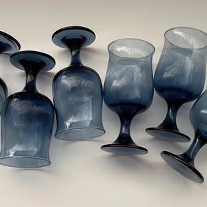 May include: Six blue-tinted wine glasses with a classic stem design. The glasses have a rounded bowl and a dark blue base and stem. The glasses are arranged on a white surface, showcasing their elegant shape and color.
