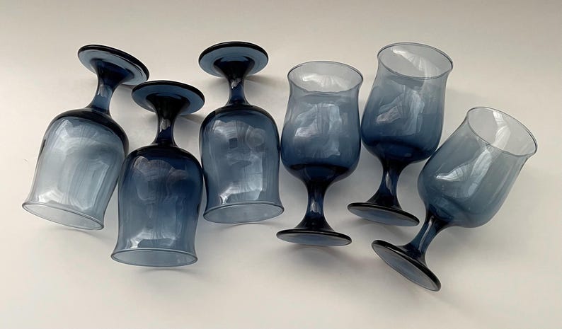 May include: Six blue-tinted wine glasses with a classic stem design. The glasses have a rounded bowl and a dark blue base and stem. The glasses are arranged on a white surface, showcasing their elegant shape and color.