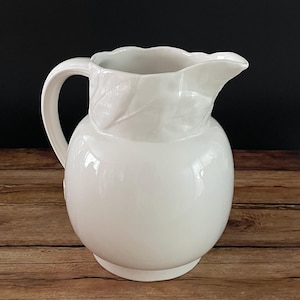 May include: A white ceramic pitcher with a rounded body, a curved handle, and a spout. The pitcher has a leaf design near the spout and a scalloped rim. It sits on a wooden surface against a dark background.