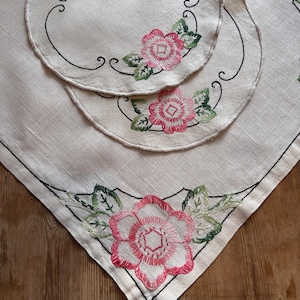 May include: Cream-colored linen table setting with embroidered pink roses and green leaves. The set includes a square tablecloth and two round placemats, each with a black border and floral design. The table setting is on a wooden surface.