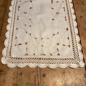 May include: A rectangular off-white table runner featuring a central solid fabric panel surrounded by intricate cutwork embroidery depicting large floral motifs. The edges are finished with a decorative scalloped crochet lace border.