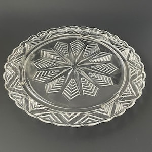 May include: A clear glass cake plate with a decorative, starburst pattern. The plate has a scalloped edge.