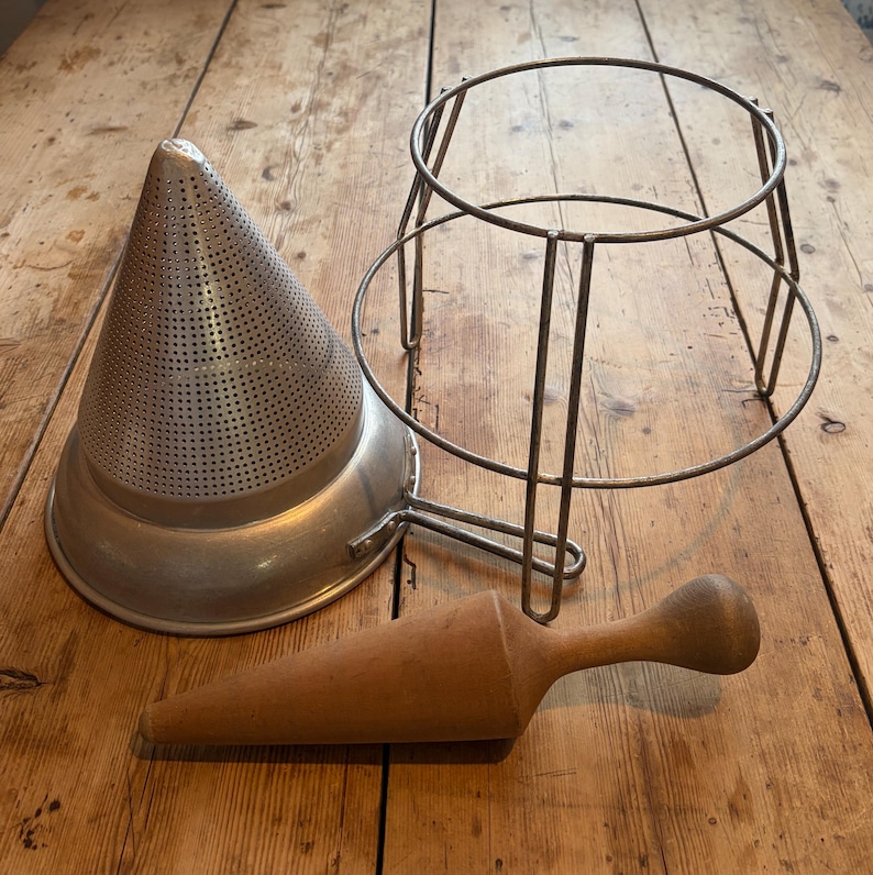 May include: A silver metal conical food mill strainer with a perforated surface, a matching metal wire stand with a circular base, and a smooth, light brown wooden pestle with a tapered conical shape and a rounded handle end.