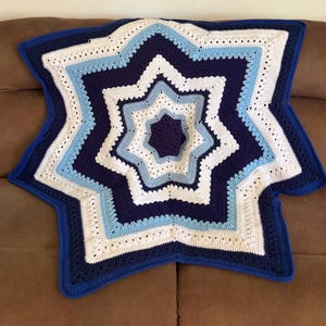 May include: A star-shaped crochet blanket featuring concentric layers of yarn in shades of dark navy blue, light sky blue, and crisp white. The design consists of eight points radiating from a central point, with a textured stitch pattern.