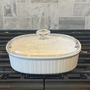 May include: White oval baking dish with a glass lid on a black stovetop. The dish has a fluted design and is sitting on a burner.