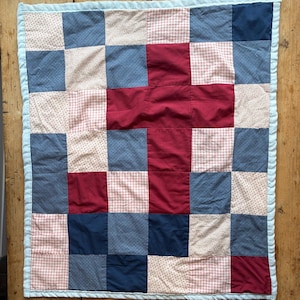 May include: A square patchwork quilt with a light blue border. The quilt features a grid pattern of red, navy blue, and light pink squares. The center of the quilt forms a red cross shape.
