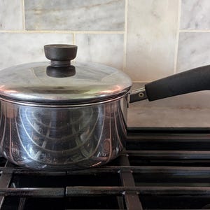 May include: A stainless steel saucepan with a lid and a black handle. The pot is sitting on a black stovetop. The lid has a black knob. The handle has a metal ring at the end.