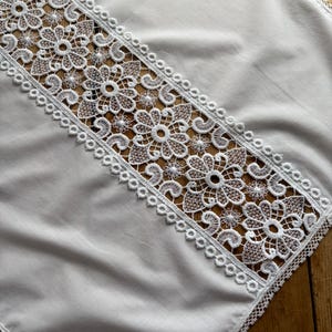 May include: A white, square tablecloth with a detailed, floral lace design running diagonally across the center. The lace features openwork flowers and a scalloped edge, adding a delicate touch to the fabric. The tablecloth is made of a soft, white material.