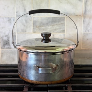 May include: A stainless steel cooking pot with a lid and a black handle. The pot has a silver finish and a copper-colored base. The lid has a dark wooden knob and the pot has a metal handle.