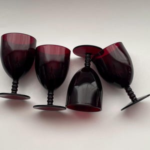 May include: Four dark red wine glasses with black stems and bases. The glasses are arranged in a semi-circle on a white surface.