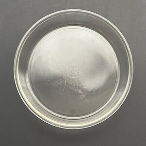 May include: A clear glass pie plate with a raised lip around the edge. The plate has a small, round, silver sticker on the bottom.