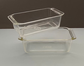 May include: Two clear glass loaf pans stacked on top of each other. Each rectangular pan has a slightly flared rim and handles on each end. The pans are made of clear glass and are on a light gray surface.