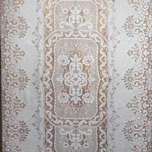 May include: White lace tablecloth with an intricate floral design. The tablecloth features a central rectangular panel with detailed floral patterns and a delicate border. The lace is set against a contrasting brown background, highlighting the design.