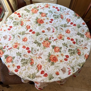 May include: A round tablecloth featuring a white background with a repeating botanical pattern of orange chrysanthemums, white flowers, green foliage, and small red berries. The fabric design covers the entire surface of the circular table.