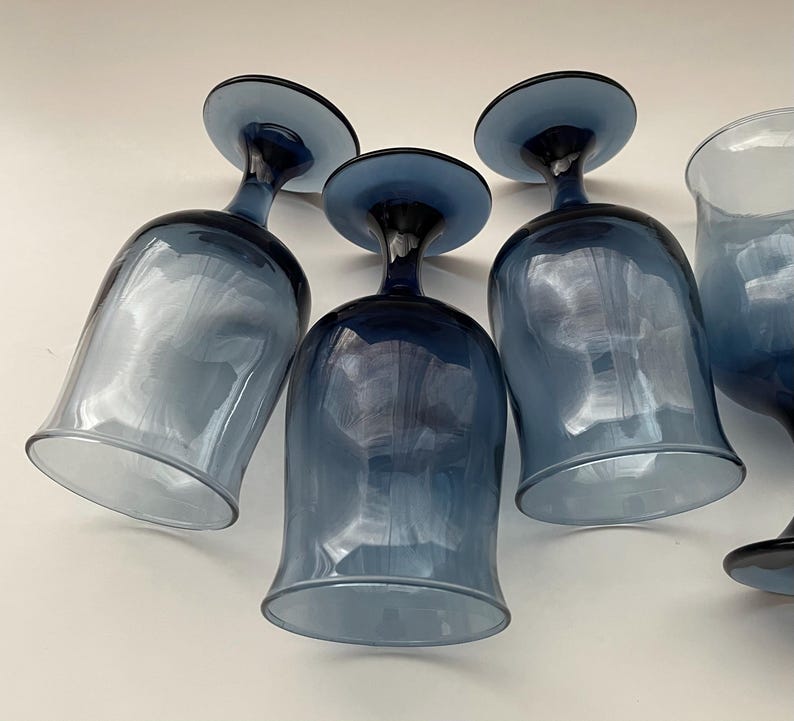 May include: A collection of blue-tinted glass wine glasses. The glasses have a rounded bowl shape and a dark blue stem and base. The glass has a gradient effect, transitioning from a lighter blue at the base to a darker blue at the stem.