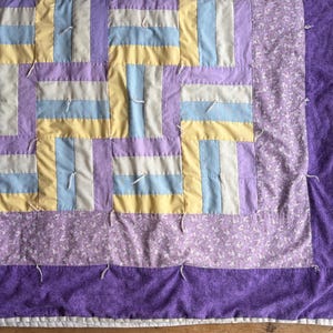 May include: A handmade quilt with a patchwork design. The quilt features a pattern of rectangular blocks in pastel colors, including lavender, yellow, and light blue. The border is a solid purple fabric. White thread is used to secure the layers.